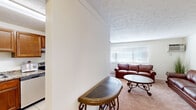 This single-floor, one-bedroom, one-bathroom residence offers a practical and comfortable layout across a compact yet efficient footprint. The combined living and dining area serves as the heart of the home, featuring soft wall-to-wall carpeting, a textured ceiling, and a wide window fitted with horizontal blinds that allows natural light to fill the space. A wall-mounted air conditioning unit provides climate control directly in the living area. The dining space accommodates a round wooden table with ample seating, seamlessly connected to the living room for easy everyday use.

The kitchen is a functional standout, featuring rich warm-toned cabinetry, granite-style countertops, light tile flooring, and stainless steel appliances including a full-size refrigerator, an electric range, and a sink with ample counter space on either side — well-suited for cooking and meal preparation.

The bedroom is a generously proportioned retreat with soft carpeting, a ceiling fan, and a louvered closet door providing convenient storage access.

The bathroom is clean and well-appointed, featuring tile flooring, white cabinetry with an integrated vanity, a mirrored medicine cabinet with overhead lighting, white tile wainscoting, and a shower/tub combination. A connecting hallway links all areas of the home efficiently, completing this thoughtfully arranged single-level residence.