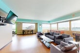 This generously sized single-floor space combines a fully equipped kitchen with a expansive open-plan living and lounge area, creating a versatile communal environment. The interior is defined by bold, energetic color choices — vivid teal walls and ceilings contrast with warm amber and sage green tones across distinct zones, giving each area a unique character while maintaining visual cohesion throughout.

The kitchen is a functional centerpiece, featuring rich dark wood cabinetry, dark granite countertops, stainless steel appliances, and a large island finished in a striking teal hue with seating for multiple occupants along its bar-height counter. Pendant lighting adds warmth overhead, and a secondary kitchenette with additional cabinetry and appliances offers supplementary functionality.

The living area is expansive and open, with hardwood-style flooring running throughout. Large horizontal windows line the walls, flooding the space with natural light and offering elevated panoramic views of the surrounding cityscape. Multiple seating arrangements accommodate different uses, from casual lounging on gray upholstered sofas and armchairs to recreation facilitated by foosball tables. A wall-mounted flat-screen television, decorative sconce lighting, and a quieter seating alcove with sage green walls round out the amenities, making this a well-appointed, multi-functional gathering space.