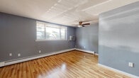 This single-floor unit is thoughtfully laid out across several rooms, each finished with consistent cool gray-blue walls and crisp white trim that create a calm, cohesive atmosphere throughout. The main living area is generously sized, featuring warm-toned hardwood flooring that gleams under natural light from a large window fitted with horizontal blinds. A ceiling fan with a light fixture is centrally mounted, while a wall-mounted air conditioning unit and perimeter baseboard heating ensure year-round comfort. The space also includes a dedicated entry door and closet access along one wall.

The bedroom features lighter vinyl plank flooring with a wood-look finish, a built-in closet, a window with outdoor views, and its own baseboard heater, offering a quiet and functional retreat.

The galley-style kitchen is efficiently designed with rich cherry-toned wood cabinetry, light stone-finish countertops, a mosaic tile backsplash, and white appliances including a gas range, microwave, and refrigerator. Tile flooring completes the functional workspace.

The bathroom features white subway tile wainscoting, a blue-gray upper wall, tile flooring, and a baseboard heater, maintaining the unit's consistent aesthetic throughout.