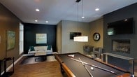 This single-floor space is designed for both recreation and relaxation, combining a well-appointed game area with a comfortable lounge zone. At the heart of the room sits a full-sized pool table with a rich wood frame, complemented by a sleek rectangular pendant light suspended overhead that draws the eye upward and anchors the space. Recessed ceiling lighting provides consistent, even illumination throughout.

The lounge area is defined by a neutral-toned sofa accented with teal throw pillows, paired with a coffee table atop a textured area rug. A bold dark accent wall serves as a backdrop for a framed abstract artwork, adding a striking visual contrast to the warmer, earthy tones found elsewhere in the room. A wall-mounted flat-screen television is positioned for easy viewing from multiple areas.

Adjacent seating includes two upholstered grey chairs near a decorative round mirror, offering additional gathering options. The warm hardwood flooring ties the entire space together, lending a grounded, sophisticated feel. Overall, the space strikes a balance between entertainment functionality and contemporary interior design.