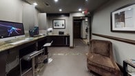 This hotel amenity space serves as a combined business center and lounge, thoughtfully designed to accommodate guests' work and leisure needs in a single, cohesive environment. The room features neutral-toned walls accented by dark wood trim and recessed lighting overhead, creating a polished and professional atmosphere throughout.

The business center area is equipped with multiple desktop computer workstations lined along a granite-topped counter with privacy dividers, accompanied by modern adjustable bar-height stools. The counter wraps along the perimeter, maximizing workstation capacity while maintaining an open feel in the center of the room.

A dedicated refreshment station occupies one corner, offering coffee and beverage equipment alongside cabinetry with frosted-glass panel doors and a granite countertop. Decorative touches, including a framed vintage espresso advertisement and a collection of curated art prints, add character and warmth to the functional space.

The lounge area features plush upholstered armchairs arranged around a small magazine rack, providing a comfortable spot for guests to relax or browse reading materials. Framed café-themed artwork adorns the walls, reinforcing the inviting ambiance. Large-format tile flooring with inlaid stone border detailing ties the space together, while ample recessed lighting ensures the entire area is well-lit and welcoming.