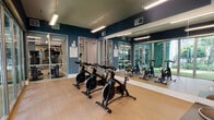 This spin studio is thoughtfully designed to deliver an immersive and motivating cycling experience. The room is finished with bold deep-blue accent walls trimmed in white, creating a sleek and energizing atmosphere. Light wood-look flooring runs throughout, lending warmth to the space while remaining practical for fitness use. A row of Schwinn stationary cycling bikes is arranged to give each rider ample space, all oriented toward a large wall-mounted flat-screen TV ideal for guided workouts or on-demand fitness content. A secondary screen is also positioned on the wall for additional viewing options. Full-length mirrors line one wall, visually expanding the room and allowing cyclists to monitor their form during sessions. The standout architectural feature is the expansive floor-to-ceiling glass wall fitted with wide sliding panel doors, which floods the studio with abundant natural light and offers open views of a lush, landscaped outdoor area with tropical palms and greenery. This seamless indoor-outdoor connection creates an airy, resort-like feel. Overhead linear LED lighting provides consistent illumination throughout. An adjacent room, visible through glass partitions, appears to house additional fitness equipment, suggesting this spin studio is part of a broader fitness facility.
