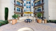This residential complex is designed around a welcoming central courtyard that serves as the heart of the community. The outdoor gathering space features a herringbone-patterned brick paver patio with a picnic table shaded by a yellow market umbrella, creating a functional and inviting communal area. Large terracotta planters with mature trees flank the space, adding warmth and greenery to the setting.

The building's exterior combines smooth stucco facades with detailed stacked-stone accents, giving it a Southwestern architectural character. Decorative stone archways with rounded corbels frame the property's entry and transition areas, while a wood-beam pergola provides partial overhead coverage near the gated entrance. Wrought-iron fencing and gates define the perimeter, contributing to a secure and polished aesthetic.

The multi-story structure features individual balconies with dark metal railings on each level, offering residents private outdoor space. Ground-floor units are accessed through six-panel metal doors set within stucco-framed entryways. Landscaped beds with low ground cover and shrubs line walkways, complementing the overall design. The combination of natural materials, detailed stonework, and open-air amenities creates a cohesive and community-oriented living environment.