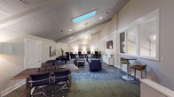 This well-appointed single-floor space is designed to accommodate a variety of functions within an open, airy layout. The most striking architectural feature is the vaulted, beadboard-paneled ceiling, which rises dramatically overhead and is punctuated by multiple skylights that flood the interior with natural light. Woven pendant light fixtures in organic, sculptural forms hang at varying heights, adding warmth and visual interest throughout.

The space is organized into several distinct functional zones. A central lounge area features tufted gray sofas, upholstered armchairs, and a low coffee table arranged over a richly patterned blue-and-green carpet, creating a comfortable gathering environment. Adjacent to this is a dedicated workstation area, where a row of computer stations lines a shiplap-accented wall beneath warm task lighting—ideal for productivity. A wall-mounted flat-screen television is positioned above a white console cabinet, anchoring one side of the lounge.

A bar-height table and stools offer an additional seating option near interior windows with frosted glass panels. Toward the entry, a refreshment nook features a professional coffee machine, cabinetry, and decorative hexagonal mirrors. A multifunction printer and wood-topped credenza round out the practical amenities, with abstract artwork and potted plants adding curated finishing touches throughout.