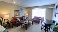 This single-floor residence offers a thoughtfully designed living space that combines comfort and functionality. The living room serves as the heart of the home, featuring plush wall-to-wall carpeting in a warm neutral tone that adds a cozy feel underfoot. Large vertical-blind-covered windows allow an abundance of natural light to fill the room, creating a bright and airy atmosphere. The space is generously proportioned, easily accommodating a full sectional sofa, an oversized accent chair with ottoman, and a glass-topped coffee table — all contributing to an inviting and relaxed setting. Walls are painted in soft, neutral tones that complement the rich, earthy upholstery throughout. A textured ceiling adds subtle architectural character, while track lighting provides focused illumination. Decorative wall art and a classic wall clock add personality to the space without overwhelming it. Climate control is well-served by ceiling vents positioned throughout the room. The large window at the far end hints at an outdoor access point, bringing the sense of openness and connectivity to the exterior. Overall, the space presents a clean, comfortable, and functional living environment well-suited for everyday living.