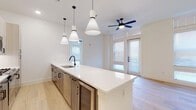 This single-floor unit showcases a clean, contemporary design throughout. The open-concept kitchen and living area serves as the heart of the space, anchored by a generously sized white quartz island with an undermount sink and matte black faucet. Three cone-shaped pendant lights hang above the island, while light wood cabinetry, a subway tile backsplash, and stainless steel appliances — including a refrigerator, microwave, and wine cooler — complete the kitchen. Light hardwood flooring flows seamlessly from the kitchen through the living area, which benefits from abundant natural light through tall windows and a glass-paneled door. A distinctive ceiling fan with blue blades adds a modern accent overhead.

The bedrooms continue the light, airy aesthetic with large windows, ceiling fans, and the same hardwood flooring. One bedroom features a glass door leading to an exterior exit. Two well-appointed bathrooms round out the unit: one features a glass-enclosed walk-in shower with marble-style tile surround and a sleek vanity with matte black hardware, while the second offers a soaking tub with a matching marble-tile surround. Both bathrooms feature large-format tile flooring and white quartz countertops, maintaining a cohesive, upscale finish throughout.