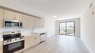 This contemporary 2-bedroom, 1-bathroom unit offers a thoughtfully designed open-concept layout with a clean, modern aesthetic throughout. The combined kitchen and living area serves as the heart of the unit, featuring light wood laminate flooring, crisp white walls, and abundant natural light. The kitchen is well-appointed with light wood cabinetry, white quartz countertops, a white tile backsplash, and a full suite of stainless steel appliances including a refrigerator, range, microwave, and dishwasher. The living area flows seamlessly toward a large sliding glass door that opens to a private balcony, providing open outdoor views and filling the space with natural light. Both bedrooms are finished with the same light wood flooring and neutral tones, with the primary bedroom featuring a large sliding glass door balcony access and the secondary bedroom offering a black-framed window with exterior views. The bathroom is elegantly finished with marble-style tile flooring, a light wood vanity with white countertop, framed mirrors, and a marble-tiled tub and shower surround. A hallway connects the main living areas to the bedrooms and bathroom, with an entrance that includes a utility closet and electrical panel access, completing this efficiently designed and stylishly finished unit.