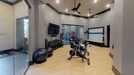 Located in Thompson's Station, TN, this single-level fitness center at 2306 Vintage Tollgate Dr is thoughtfully designed to support a wide range of workout routines. The space features light hardwood-style flooring throughout, neutral gray walls accented with dark trim, and elevated ceilings that lend an open, airy atmosphere. Ceiling fans are strategically positioned to maintain airflow during workouts.

The main exercise area houses multiple spin bikes arranged for group cycling sessions, accompanied by a wall-mounted flat-screen television and an interactive display panel for guided workouts. A well-stocked free weights station features an organized rack of kettlebells and a colorful assortment of medicine balls in varying weights, alongside stability and exercise balls for functional training.

A separate strength training zone is enclosed by full-length glass walls, offering visibility while maintaining a distinct workout environment. Inside, cable machines, a weight bench, and additional strength equipment are arranged efficiently. A large mirror along the wall enhances the sense of space. Near the entrance, a built-in storage bench with cubbies and a decorative herringbone accent panel provide a practical and stylish transition area. Recessed lighting throughout ensures a bright, welcoming environment conducive to focused exercise.