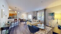 Located at 4 Seven Springs Lane in Burlington, Massachusetts, this single-floor residence offers a thoughtfully designed open-concept layout that seamlessly connects the living, dining, and kitchen areas. The living room is a bright and welcoming space, featuring large windows dressed with floor-length curtains that invite generous natural light and views of the surrounding greenery. Wood-look plank flooring flows throughout the main living areas, adding warmth and continuity to the design.

The kitchen is well-equipped with rich warm-toned cabinetry, dark granite countertops, a subway tile backsplash, and a full suite of stainless steel appliances including a range, microwave, refrigerator, and dishwasher. A breakfast bar provides additional seating and connects the kitchen to the dining area.

The bedroom is a serene retreat, featuring plush carpeting, large windows with outdoor views, and ample space for a full furniture arrangement. The bathroom complements the overall aesthetic with matching warm wood cabinetry, dark granite countertops, a backlit vanity mirror, and a full tub-shower combination.

A hallway connects the living spaces to the private areas of the residence, with additional storage closets along the way. The consistent use of neutral tones, modern fixtures, and quality finishes throughout creates a cohesive and comfortable living environment.