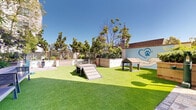 Located at 98 N 1st St in downtown San Jose, California, this thoughtfully designed outdoor dog park offers a dedicated, pet-friendly retreat surrounded by a modern residential community. The space features a generous expanse of lush artificial turf, providing a clean and comfortable surface for dogs to roam, play, and socialize year-round. Agility equipment, including ramps and a small doghouse structure, is strategically placed throughout the area to encourage active play and enrichment. A classic red fire hydrant adds a playful, iconic touch to the landscape. Raised wooden planter beds line portions of the perimeter, filled with greenery and shrubs that contribute to a welcoming, park-like atmosphere. Seating benches are positioned along the edges, offering pet owners a comfortable place to relax while supervising their animals. A pet wash station and water access point are also present, ensuring convenience for both pets and their owners. The space is enclosed by low retaining walls and bordered by mature trees, providing shade and a sense of seclusion within the urban environment. A decorative mural featuring a paw print motif adds a charming, community-oriented character to the space. Abundant natural sunlight floods the area, enhancing the open-air ambiance of this well-appointed pet amenity space.