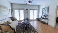 This well-appointed single-floor space exudes a warm and inviting atmosphere, thoughtfully designed with a blend of classic elegance and comfortable living. The living area is a standout feature, showcasing rich hardwood flooring that extends throughout and serves as a grounding element for the refined furnishings. A ceiling fan with an integrated light fixture is centrally positioned overhead, providing both comfort and ambient lighting.

Natural light pours generously into the room through multiple large French doors fitted with white plantation shutters, creating a bright, airy feel while maintaining privacy. Neutral-toned walls complement the light-filled environment and serve as a versatile backdrop for the décor.

The living area is anchored by a patterned area rug in deep, earthy tones, over which sits a circular glass-top coffee table with ornate dark wood legs. A plush sofa with decorative pillows adds to the room's comfortable and classic aesthetic. Along the far wall, a slim console table with decorative accents and an accent bench provide additional style and function. Wall art, including a framed landscape piece, adds personality and visual interest to the space.

Overall, the space presents a cohesive, traditionally styled interior that balances elegance with everyday livability.