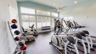 Located at 10765 US-78 E in Summerville, SC, this fitness center is designed to support a comprehensive workout experience within a single-floor layout. The space is finished with soft aqua-toned walls and durable rubber flooring, creating a clean, functional environment suited for regular exercise use.

The main workout area is generously sized and bathed in natural light, courtesy of a large bank of windows along the exterior wall fitted with adjustable blinds. Vaulted ceilings with recessed lighting and a ceiling fan enhance the open, airy feel of the space while supporting airflow and comfort during workouts.

Cardio equipment is well-represented throughout the facility, including multiple treadmills, elliptical trainers, stationary upright bikes, and a recumbent bike. A dedicated cable and pulley strength machine provides options for resistance training, while a full rack of dumbbells offers versatility for free weight exercises. A medicine ball storage tower with multiple weighted balls rounds out the functional fitness offerings.

The layout is thoughtfully organized, with cardio machines positioned to face the large windows, providing pleasant outdoor views of the surrounding greenery. The overall ambiance is bright, welcoming, and professionally appointed, making this fitness center a practical and well-maintained amenity for the Summerville community.