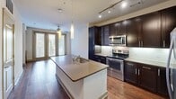 This well-appointed 2-bedroom, 1-bathroom residence in Dallas, Texas offers a thoughtfully designed single-floor layout. The open-concept kitchen serves as the heart of the home, showcasing rich dark-espresso cabinetry, stainless steel appliances including a range and microwave, a tile backsplash, and a large central island with a sink and pendant lighting overhead. Track lighting and hardwood flooring extend through the main living areas, adding warmth and a modern touch. French doors allow natural light to pour into the living space. The primary bedroom is generously sized, featuring plush carpeting and a ceiling fan, with direct access to the bathroom and a dedicated closet. The second bedroom also features hardwood floors, a ceiling fan, and a built-in closet. The bathroom is finished with large-format tile flooring, a dark-cabinet vanity with a framed mirror, and ample drawer storage. A dedicated laundry area with shelving, washer/dryer hookups, and tile flooring provides added convenience. Neutral wall tones, panel-style doors, and consistent finishes throughout give the space a cohesive and clean aesthetic.