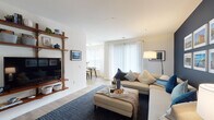 This thoughtfully designed single-floor residence offers a bright, open-concept layout that seamlessly connects the living, dining, and kitchen areas. The living room is a welcoming space anchored by wood-toned freestanding shelving that houses a large flat-screen TV, complemented by plush seating and light hardwood-style flooring. Sheer white curtains and vertical blinds allow abundant natural light to filter throughout. The adjacent dining area is framed by large corner windows, creating a sun-filled nook ideal for gathering.

The kitchen features gray shaker-style cabinetry, granite-look countertops, and black appliances including a gas range, microwave, and refrigerator, all set over continued hardwood-style flooring. A hallway with built-in coat hooks and wall-mounted mirror leads to the private quarters. Both bedrooms offer plush carpeting and bold dark accent walls that create a cozy, sophisticated atmosphere, with ample space for a desk or dresser. The bathroom is finished with a granite-topped vanity, wood-toned flooring, and decorative geometric shelving. A dedicated in-unit laundry area with side-by-side washer and dryer and overhead shelving completes this functional and stylish residence.