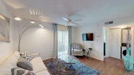 This single-floor residence offers a well-organized layout that balances comfort and functionality across its living spaces. The living room serves as the central gathering area, finished with warm hardwood-style flooring and anchored by a ceiling fan overhead. Natural light filters in through a sliding glass door dressed with teal patterned drape panels and vertical blinds, while a wall-mounted flat-screen television and arc floor lamp complete the space. A compact dining area sits adjacent, featuring a round glass-top table with white chairs and a large wall mirror that visually expands the room. A pass-through window connects the dining area to the kitchen, which is equipped with granite-style countertops, white appliances including a full-size refrigerator, and warm wood cabinetry. The bedroom offers plush carpeting, a ceiling fan, a wall-mounted television, and a generously sized window that invites natural light. The bathroom is cleanly finished with tile flooring, wainscoting, a granite-topped vanity, and a shower-tub combination. Throughout the residence, a consistent neutral color palette with teal and grey accents ties the spaces together, creating a cohesive and inviting ambiance.