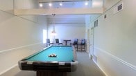 This single-floor recreation room is thoughtfully designed to serve as a social and leisure hub. The centerpiece of the space is a full-size pool table with a classic green felt surface, positioned prominently beneath a stylish pendant light fixture that provides focused illumination for gameplay. The high ceiling with an elevated upper ledge adds a sense of openness and volume to the room, enhancing its spacious feel.

Adjacent to the pool table is a casual dining and lounge area, furnished with a round high-top table and upholstered bar-height chairs in dark tones, creating an inviting spot for conversation or refreshments. The neutral color palette of white and beige walls, complemented by light-toned carpeting, gives the room a clean and versatile aesthetic.

Recessed track lighting along the upper ceiling level ensures the entire space is well-lit, while a window on the far wall allows for natural light to filter in. Built-in HVAC vents are integrated seamlessly into the walls, ensuring year-round comfort. A doorway at the rear of the room provides easy access to adjoining areas. Overall, this recreation room offers a well-appointed environment ideal for casual entertainment and community gatherings.
