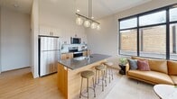 Located at 375 W Whitney Ave in Salt Lake City, Utah, this thoughtfully designed one-bedroom, three-bathroom residence offers a contemporary living experience within a compact yet efficient single-floor layout. The open-concept living area seamlessly integrates the kitchen, dining, and living spaces, creating an inviting and functional hub for daily life. The modern kitchen is a standout, featuring a large central island with a quartz countertop and bar seating, warm wood cabinetry paired with white upper cabinets, a white subway tile backsplash, and stainless steel appliances. A striking linear pendant light with globe fixtures illuminates the island and adjoining living space. Large black-framed windows flood the living area with natural light and offer views of the surrounding neighborhood. The bedroom is a calm retreat, appointed with soft neutral tones, plush carpet, a built-in closet with sliding panel doors, and exposed concrete architectural accents. Three well-appointed bathrooms include a full bath with a tub-shower combination and a primary bath featuring warm wood vanity cabinetry and modern fixtures. Light wood-tone flooring flows throughout the main living areas, complemented by clean white walls and black hardware details. A dedicated laundry area and multiple closets add to the unit's practicality.