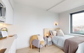 This thoughtfully designed studio unit occupies a single floor and seamlessly integrates living, sleeping, and cooking functions within a bright, open-plan layout. The interior is finished in a clean, contemporary palette of white and soft neutrals, complemented throughout by light-toned hardwood flooring that enhances the sense of openness and warmth.

The sleeping area serves as the focal point of the main space, anchored by a bed with a natural wood headboard and flanked by a wall-mounted sconce that provides warm ambient lighting. A compact lounge chair adds a cozy, functional corner for relaxing. A large picture window floods the room with natural light and offers expansive city views, making the space feel airy and connected to its urban surroundings.

The kitchenette is fully integrated along one wall, featuring sleek white flat-front cabinetry extending to the ceiling, an induction cooktop, an over-the-counter microwave, and a sink — providing all essential cooking and storage amenities in an efficient, streamlined design. A dining area with a small table and chair is positioned nearby, separated from the main room by a floor-to-ceiling curtain panel.

The bathroom is finished with cool grey walls and tiled flooring, housing a wall-mounted toilet, a recessed storage niche, and a large mirror, delivering a clean and functional space with a modern aesthetic.