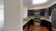 This single-floor residence offers a thoughtfully designed layout that balances style and functionality throughout. The kitchen stands out as a centerpiece, showcasing dark espresso cabinetry with beadboard detailing, granite countertops, a tile mosaic backsplash, and a full suite of stainless steel appliances including a range, over-the-range microwave, dishwasher, and refrigerator. Rich hardwood flooring flows seamlessly from the kitchen into the open living and dining areas, creating a warm and cohesive aesthetic. The spacious living room is bathed in natural light from large windows and sliding glass doors that lead to a private balcony, while a built-in electric fireplace serves as a focal point along the wall. The bathroom features a dual-sink vanity with matching granite countertops, dark cabinetry, a large mirror, and contemporary wall sconces set against elegant vertical tile. Multiple walk-in closets with wire shelving systems provide generous storage throughout the residence. A dedicated laundry room accommodates a full-sized top-load washer and dryer, adding everyday convenience. Neutral warm-toned walls and clean white trim complement the overall design, creating a polished and inviting atmosphere.
