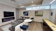 This contemporary single-floor lounge is thoughtfully designed with a clean, modern aesthetic. The space features smooth neutral-toned walls complemented by warm wood-look flooring, creating a welcoming and polished atmosphere throughout. A striking stacked-stone fireplace with a linear electric insert serves as a focal point on one wall, adding both warmth and visual interest to the room.

Multiple wall-mounted flat-screen displays are positioned throughout the space, making it well-suited for entertainment or collaborative gatherings. Two pieces of framed art photography — featuring vivid macro imagery — add color and personality to the neutral walls. High-positioned clerestory windows run along one wall, drawing in natural light while maintaining privacy at floor level.

Seating is arranged to encourage social interaction, with a large sectional sofa in charcoal gray anchoring the main lounge area, accompanied by a low coffee table. A pair of round white high-top tables with sleek chrome bases and deep navy blue upholstered bar stools provide a casual setting for smaller gatherings. Recessed ceiling lighting offers even, adjustable illumination throughout. The overall ambiance is modern, refined, and versatile — ideal for relaxing, entertaining, or informal meetings.
