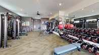Located in Camp Verde, Arizona, this fitness center offers a generously sized, single-floor workout facility designed to support a full range of exercise routines. The space features a neutral-toned geometric carpet floor and recessed lighting throughout, complemented by ceiling fans that help maintain a comfortable environment during workouts. Large floor-to-ceiling mirrors line the walls, enhancing the sense of openness and allowing users to monitor their form. A distinctive architectural feature is the glass-panel garage-style door that opens to the outdoors, flooding the space with natural light and providing views of the surrounding exterior area.

The facility is comprehensively equipped with a wide array of cardio machines, including treadmills, ellipticals, a stair climber, and stationary bikes, many outfitted with individual display screens. The strength training section includes a multi-station cable machine, a leg press, adjustable weight benches, and a full rack of dumbbells in varying weights. Stability balls and medicine balls are neatly organized along the mirrored wall, adding versatility for functional training. Wall-mounted flat-screen televisions are positioned throughout the space for entertainment. The overall ambiance is modern, clean, and functional, making it a well-rounded fitness destination.