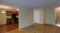 This single-floor residence offers a straightforward and functional layout designed for comfortable everyday living. The open living area is finished with wall-to-wall carpeting in a neutral tone, creating a warm and inviting atmosphere. Soft, light-colored walls throughout contribute to a clean and airy feel.

Adjacent to the living area, the kitchen is defined by rich wood cabinetry in a warm brown finish, providing ample storage space. The kitchen is equipped with stainless steel appliances, including a range, and features dark countertops that complement the cabinetry. The kitchen floor transitions to hardwood-style flooring, offering a practical and durable surface in the cooking area.

Interior doors throughout the space are finished in classic white with a raised four-panel design, adding a touch of traditional character. Hardware includes lever-style handles for a modern, accessible touch.

The layout benefits from a clear separation between the kitchen and living spaces, defined by a partial wall that provides distinction between areas while maintaining an open feel. Overall, the space presents a clean, move-in-ready environment with a cohesive neutral palette and a mix of practical finishes suited for a variety of lifestyles.