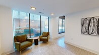 Located at 142 W York St in Norfolk, Virginia, this single-floor space delivers a refined, contemporary aesthetic defined by clean lines and an abundance of natural light. Floor-to-ceiling windows along the street-facing walls flood the interior with daylight while offering open views of the surrounding urban streetscape, creating an inviting connection between indoors and out.

The layout flows between distinct functional zones. A seating lounge area features light wood-toned flooring, neutral area rugs, and a curated mix of seating — including plush cream barrel chairs and olive green mid-century armchairs — accented by tasteful wall art and greenery. The space transitions into a well-appointed kitchen area anchored by a generously sized island with a dramatic dark marble-veined countertop, deep green cabinetry, and gold-accented brass fixtures. Stainless steel appliances, a tiled backsplash in coordinating tones, and pendant lighting complete the kitchen's polished look.

Soaring ceiling heights lend an open, airy quality throughout, reinforcing the sense of spaciousness. The neutral white walls serve as a versatile backdrop, complementing both the warm wood flooring and the bold design accents. Overall, the space presents a sophisticated, modern environment well-suited for a variety of uses in the heart of downtown Norfolk.