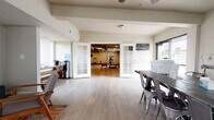 This well-appointed single-floor space offers a thoughtfully organized layout designed for both functionality and comfort. The main entry area features light wood-tone flooring, a work table with industrial-style metal chairs, and direct access to an outdoor area, making it ideal as a reception or administrative zone. A pair of elegant French doors with frosted glass panels connects this area to the interior, lending a polished, professional feel to the transition between spaces.

The central lounge area is warm and inviting, furnished with mid-century modern accent chairs, a colorful area rug, and a wall-mounted television, creating a comfortable gathering spot. Natural light filters in through multiple windows, brightening the neutral-toned walls throughout.

Beyond the French doors lies a spacious fitness area, equipped with a multi-station weight machine, a treadmill, a stationary bike, TRX suspension straps, and wall-mounted televisions. The fitness zone features a mix of hardwood-style flooring and carpet tile, with recessed lighting and a ceiling fan providing a well-lit, comfortable environment for exercise.

Additional open rooms with light laminate flooring, recessed lighting, and textured ceilings provide flexible space for meetings, events, or other uses. A hallway with mirrors and seating benches serves as a transitional zone, completing this multi-purpose, move-in-ready space.