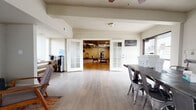 This well-appointed single-floor space offers a thoughtfully organized layout designed for both functionality and comfort. The main entry area features light wood-tone flooring, a work table with industrial-style metal chairs, and direct access to an outdoor area, making it ideal as a reception or administrative zone. A pair of elegant French doors with frosted glass panels connects this area to the interior, lending a polished, professional feel to the transition between spaces.

The central lounge area is warm and inviting, furnished with mid-century modern accent chairs, a colorful area rug, and a wall-mounted television, creating a comfortable gathering spot. Natural light filters in through multiple windows, brightening the neutral-toned walls throughout.

Beyond the French doors lies a spacious fitness area, equipped with a multi-station weight machine, a treadmill, a stationary bike, TRX suspension straps, and wall-mounted televisions. The fitness zone features a mix of hardwood-style flooring and carpet tile, with recessed lighting and a ceiling fan providing a well-lit, comfortable environment for exercise.

Additional open rooms with light laminate flooring, recessed lighting, and textured ceilings provide flexible space for meetings, events, or other uses. A hallway with mirrors and seating benches serves as a transitional zone, completing this multi-purpose, move-in-ready space.