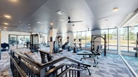 Located at 100 Waddell Plaza Lane in Cary, NC, this modern fitness facility spans a single floor and offers a comprehensive workout environment across two distinct exercise rooms. The main gym area features patterned, multi-tone carpet tiles and recessed lighting set within a sleek dark ceiling, creating a focused and energetic atmosphere. This space is outfitted with an extensive array of cardio equipment — including treadmills, ellipticals, and cable machines — along with free weights, adjustable benches, and functional strength stations. Floor-to-ceiling mirrors line one wall, visually expanding the space and allowing users to monitor their form. Large windows along the perimeter invite abundant natural light and offer views of the surrounding outdoors. The second exercise room is designed for functional and group training, featuring wide-plank hardwood flooring, ceiling fans, and a wall-mounted equipment rack stocked with medicine balls, battle ropes, and kettlebells. Floor-to-ceiling glass partitions and mirrored walls with motivational text add a dynamic, studio-like feel. Rowing machines and Pilates reformers are also present, catering to a variety of fitness disciplines. Together, both rooms deliver a polished, versatile, and well-lit fitness experience.
