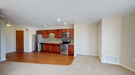 This single-floor space showcases an open-concept layout that connects a well-appointed kitchen to an adjacent living area. The kitchen is outfitted with a full suite of stainless steel appliances, including a refrigerator, electric range, built-in microwave, and dishwasher. Rich, warm-toned wood cabinetry extends along the full length of the kitchen wall, providing generous storage above and below the countertops. A mosaic tile backsplash in neutral tones adds a stylish detail between the upper and lower cabinets, complementing the overall design. Recessed lighting in the kitchen ceiling ensures bright, functional illumination throughout the cooking space. The kitchen floor features hardwood-style flooring that transitions into plush carpeting in the adjoining living area, clearly delineating the two functional zones while maintaining a cohesive aesthetic. The living area is open and airy, with neutral wall tones and standard-height ceilings that lend a clean, versatile atmosphere. A door along the far wall suggests access to additional rooms or a utility area. The overall design balances practicality with a comfortable, modern aesthetic.