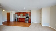 This single-floor space showcases an open-concept layout that connects a well-appointed kitchen to an adjacent living area. The kitchen is outfitted with a full suite of stainless steel appliances, including a refrigerator, electric range, built-in microwave, and dishwasher. Rich, warm-toned wood cabinetry extends along the full length of the kitchen wall, providing generous storage above and below the countertops. A mosaic tile backsplash in neutral tones adds a stylish detail between the upper and lower cabinets, complementing the overall design. Recessed lighting in the kitchen ceiling ensures bright, functional illumination throughout the cooking space. The kitchen floor features hardwood-style flooring that transitions into plush carpeting in the adjoining living area, clearly delineating the two functional zones while maintaining a cohesive aesthetic. The living area is open and airy, with neutral wall tones and standard-height ceilings that lend a clean, versatile atmosphere. A door along the far wall suggests access to additional rooms or a utility area. The overall design balances practicality with a comfortable, modern aesthetic.