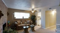 This single-floor residence offers a warm and comfortable layout designed for easy everyday living. The living room serves as the heart of the space, featuring wall-to-wall carpeting in a neutral tone that adds softness underfoot. A ceiling fan with a built-in light fixture is centrally mounted on the textured popcorn ceiling, providing both airflow and ambient lighting. A large window fitted with white horizontal blinds allows natural light to fill the room, brightening the warm taupe-toned walls. The living area comfortably accommodates seating and accent furniture, creating an inviting atmosphere for relaxing or entertaining. Decorative touches such as potted plants add a natural, lived-in charm to the space. The layout flows seamlessly into an adjacent kitchen, which is visible through an open passageway. The kitchen features white upper cabinetry, white appliances including a refrigerator and range, and a clean, functional design. The open connection between the living room and kitchen enhances the overall sense of space and makes the floor plan feel cohesive and practical. Overall, this residence presents a straightforward, comfortable living environment with a classic and understated aesthetic.