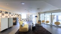 This single-floor space showcases a thoughtfully designed open-plan interior that seamlessly blends living and dining areas. The living zone is anchored by a plush, curved yellow sofa and coordinating armchair arranged atop a rich, dark shag area rug, creating a warm and inviting focal point. A sleek black coffee table serves as a functional centerpiece, accented by a sculptural decorative figure. Adjacent seating includes modern orange and grey accent chairs, adding vibrant pops of color throughout.

A striking black-and-white patterned feature wall with an animal motif provides a bold graphic contrast to the otherwise neutral palette of white ceilings and light flooring. Recessed and flush-mount ceiling lighting ensures even illumination throughout, complemented by a tall arc floor lamp that adds ambient warmth to the seating area.

The dining area features bar-height seating and a minimalist table beneath a decorative pendant light fixture, with large floor-to-ceiling windows and sliding glass doors flooding the space with natural light and providing access to an outdoor area. The overall ambiance is contemporary, curated, and sophisticated.