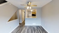 This three-floor space offers a well-organized layout designed for comfortable, everyday living. The main living area is open and airy, finished with grey-toned hardwood-style flooring that runs throughout, clean white baseboards, and soft grey walls that lend a modern, neutral aesthetic. Crown molding frames the ceiling, adding a refined architectural detail, while a ceiling fan with integrated lighting provides both air circulation and ambient illumination.

A peninsula-style kitchen opens directly to the living area, creating a seamless, connected flow between spaces. The kitchen features warm-toned cabinetry, a stainless steel refrigerator, and a countertop overhang that allows for casual seating. A second ceiling light fixture within the kitchen ensures the workspace remains well-lit.

A staircase visible from the main living area connects all three floors, suggesting a thoughtfully vertical layout with dedicated spaces spread across each level. The upper floors appear to house additional rooms, including what may be utility or storage areas, based on the structural model. Throughout the space, the consistent use of light tones and clean finishes creates a cohesive, welcoming atmosphere.