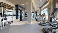 Located at 7108 E Lowry Blvd in Denver, Colorado, this contemporary fitness center is designed to deliver a comprehensive workout experience in a bright, open environment. The single-floor facility is characterized by soaring ceilings and expansive floor-to-ceiling windows that flood the space with natural light while offering panoramic views of the surrounding Denver landscape.

The cardio zone features a row of treadmills and elliptical machines positioned directly in front of the large windows, each equipped with individual screens for entertainment during workouts. Functional training is well-supported with a dedicated area featuring a heavy-duty tire sled and open rubber flooring, ideal for high-intensity training. A multi-station cable machine anchors the strength training section, complemented by a full rack of dumbbells arranged along a mirrored wall, which enhances the sense of space and allows for easy form monitoring.

A separate glass-enclosed studio offers a private space for cycling classes and group workouts, complete with its own pendant lighting and scenic outdoor views. The facility also includes a textured brick accent wall with a mounted flat-screen TV, adding a stylish, modern aesthetic. Linear pendant lighting and recessed fixtures throughout ensure the space remains well-lit at all times. The neutral color palette of warm beige and deep navy creates a focused, motivating atmosphere suited for all fitness levels.