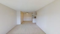 This single-floor space features an open-concept layout that seamlessly connects the living area to the kitchen. The living space is finished with wall-to-wall carpet in a neutral beige tone, creating a soft, understated foundation throughout. Smooth white walls and a white ceiling contribute to a clean, bright atmosphere, while a ceiling fan with a built-in light fixture provides both air circulation and ambient lighting to the main living area. A door near the back of the space leads to an additional room, adding functional separation within the unit. The kitchen is positioned at the rear and is defined by white cabinetry with upper and lower storage, a countertop peninsula that partially separates it from the living space, and a compact, efficient layout. The transition between the living and kitchen areas is open, enhancing the sense of flow and connectivity. Overall, the space presents a neutral, move-in-ready aesthetic with a simple and functional design that offers a blank canvas for personalization.
