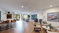 Located at 15430 Founders Lane in Apple Valley, Minnesota, this thoughtfully designed single-floor community space offers a welcoming and versatile environment for gathering, relaxing, and socializing. The open-concept layout features warm wood-tone flooring throughout, recessed lighting, and a soft, contemporary color palette of whites and muted blues that create a calm, inviting atmosphere.

The main lounge area is generously proportioned, with multiple seating arrangements that include mid-century modern accent chairs, a comfortable sofa, and marble-topped tables — ideal for both casual conversation and quiet relaxation. Large windows with warm wood-framed surrounds invite an abundance of natural light and provide views of the surrounding greenery.

A dedicated kitchenette serves the space with dark granite countertops, gray cabinetry, stainless steel appliances including a full-size refrigerator and microwave, and open wood shelving — offering convenient amenities for residents and guests alike.

The space is further enriched by curated artwork, including a striking large-scale abstract piece displayed prominently on a feature wall, alongside tasteful greenery and decorative accents. Wall sconces add a warm, layered lighting effect throughout. A secondary seating alcove provides an intimate retreat within the broader space, making this community room both functional and aesthetically refined.