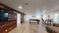 This single-floor entertainment space is thoughtfully designed to balance active recreation and relaxed lounging. The layout is anchored by a spacious game room featuring a sleek black pool table with a vivid red felt surface, positioned beneath a striking industrial-style pendant light fixture with multiple black shades. Natural light filters in through plantation shutters lining the walls, complementing the crisp white walls and light hardwood flooring that run throughout the space.

A dramatic dark wood horizontal shiplap accent wall serves as a prominent design feature, adding warmth and visual contrast against the lighter tones of the room. This same wall hosts a large flat-screen television mounted flush into the paneling, with vibrant yellow hexagonal ottomans arranged in front, creating a casual media viewing area. Framed artwork adorns sections of the accent wall, adding a curated touch.

Adjacent to the game area, a comfortable lounge zone features a large sectional sofa accented with bold red throw pillows, arranged to face the windows. A geometric black wall-mounted shelving unit and framed artwork add personality to the lounge. Recessed lighting throughout ensures consistent illumination, while a cue rack mounted near the pool table signals the space's dedication to entertainment.