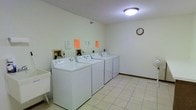This shared laundry facility is a well-organized, single-floor space designed to serve multiple users efficiently. The room features a row of top-loading washing machines paired with matching dryers, all finished in white and neatly aligned along one wall. Posted notices above the machines provide usage instructions and guidelines for users. A utility sink is situated to one side of the room, offering added convenience for hand-washing or pre-treating garments. The space is finished with light-toned ceramic tile flooring that is easy to maintain and contributes to the room's clean, bright atmosphere. White walls throughout reflect the overhead flush-mount ceiling light, ensuring the area is well-illuminated. A wall clock is mounted at eye level, allowing users to keep track of their laundry cycles with ease. The room's straightforward, functional layout maximizes usable floor space while keeping equipment accessible. Overall, this laundry facility provides a practical and tidy communal space with all the essential amenities needed for convenient, day-to-day laundry use.