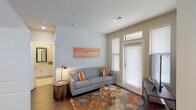 This single-story residence is thoughtfully designed with a cohesive, contemporary aesthetic that balances functionality and comfort. The living area serves as the heart of the unit, featuring rich hardwood-style flooring, neutral wall tones, and a private entry door flanked by windows fitted with slatted blinds that allow for controlled natural light. A transom window above the entry door adds an additional source of brightness to the space.

The kitchen is well-appointed with white shaker-style cabinetry extending to the ceiling, stainless steel appliances including a side-by-side refrigerator with ice maker, a built-in microwave, and a range. Light-veined granite countertops extend across the workspace and onto a peninsula with seating for multiple bar stools, making it an ideal area for casual dining or entertaining.

The bedroom offers a quieter retreat with plush carpet flooring, a ceiling fan, and a window dressed with layered blinds and sheer curtains that invite soft, natural light. The bathroom includes a deep soaking tub with tiled surround and direct access to a walk-in closet, providing practical and comfortable everyday living.