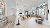 This single-floor space presents a refined, contemporary design characterized by soaring double-height ceilings and an open-plan layout that promotes a sense of expansiveness and flow. Large black-framed windows positioned at both standard and clerestory heights flood the interior with abundant natural light, offering views of surrounding greenery and creating a bright, welcoming ambiance throughout.

The main living area serves as the centerpiece of the space, anchored by a curved cream sectional sofa, organic-shaped coffee tables, and an array of seating vignettes featuring velvet accent chairs in soft blue-grey tones. A wall-mounted television is positioned within a dedicated lounge zone, complemented by sculptural pendant lighting installations that cascade dramatically from the ceiling above.

A built-in counter with warm wood cabinetry and bar-height seating defines a lounge bar area along one wall, while a dedicated workstation zone features dual monitors and ergonomic seating. An artistic textured accent wall in soft aqua tones anchors this workspace area, adding visual depth alongside curated wall art.

The hallway connects the main areas seamlessly, with sheer linen drapery, decorative lattice partitions, and light-toned large-format tile flooring completing the polished, cohesive aesthetic throughout.