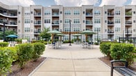 Located at 91 North York Road in Willow Grove, Pennsylvania, this outdoor courtyard space serves as the central amenity hub of a modern multi-story residential community. The building's neutral-toned vinyl siding and multi-level balconies frame the courtyard on all sides, creating a cohesive and sheltered environment that feels both open and intimate.

The courtyard is organized into several distinct zones designed for relaxation and social interaction. A spacious concrete patio at its heart features multiple seating arrangements, including white outdoor dining sets shaded by large green market umbrellas, modern pod-style lounge chairs, and striped cushioned benches. A wooden pergola serves as an architectural focal point, adding warmth and structure to the outdoor dining area. String lights are strung overhead in sections of the courtyard, lending a welcoming ambiance during evening hours.

Lush green turf lawns are enclosed by ornamental black iron fencing with decorative circular detailing, providing well-maintained green space for leisure. Adirondack chairs arranged in clusters invite casual outdoor gatherings. Manicured shrubbery and planting beds line the walkways and building perimeter, enhancing the landscape throughout. Wide concrete pathways connect all zones, ensuring ease of movement across the courtyard. The space is completed by classic lamppost lighting and thoughtfully placed benches, making it a functional and attractive outdoor retreat.