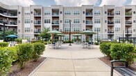 Located at 91 North York Road in Willow Grove, Pennsylvania, this outdoor courtyard space serves as the central amenity hub of a modern multi-story residential community. The building's neutral-toned vinyl siding and multi-level balconies frame the courtyard on all sides, creating a cohesive and sheltered environment that feels both open and intimate.

The courtyard is organized into several distinct zones designed for relaxation and social interaction. A spacious concrete patio at its heart features multiple seating arrangements, including white outdoor dining sets shaded by large green market umbrellas, modern pod-style lounge chairs, and striped cushioned benches. A wooden pergola serves as an architectural focal point, adding warmth and structure to the outdoor dining area. String lights are strung overhead in sections of the courtyard, lending a welcoming ambiance during evening hours.

Lush green turf lawns are enclosed by ornamental black iron fencing with decorative circular detailing, providing well-maintained green space for leisure. Adirondack chairs arranged in clusters invite casual outdoor gatherings. Manicured shrubbery and planting beds line the walkways and building perimeter, enhancing the landscape throughout. Wide concrete pathways connect all zones, ensuring ease of movement across the courtyard. The space is completed by classic lamppost lighting and thoughtfully placed benches, making it a functional and attractive outdoor retreat.