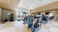Located at 1601 Maguire Rd in Ocoee, Florida, this single-floor fitness facility offers a comprehensive and thoughtfully designed workout environment. The main exercise room is expansive and airy, distinguished by soaring ceilings, warm wood-paneled walls, and a striking checkerboard-patterned floor in neutral tones. An extensive array of Matrix-brand strength machines, free weights, and cardio equipment — including treadmills, ellipticals, and air bikes — fills the space. Large floor-to-ceiling windows along the perimeter flood the room with natural light and provide views of the outdoor pool area, while ceiling fans and mounted flat-screen TVs enhance comfort. Full-length mirrors along one wall visually expand the space further. A separate studio area features light hardwood floors, a large retractable glass garage door that opens to the outdoor surroundings, and Pilates reformer equipment, making it well-suited for group fitness or individual instruction. Built-in shelving and a large wall-mounted display screen add functionality to the studio. The connecting hallways are finished with textured stone-look wall coverings, terrazzo-style flooring, and include a dual water fountain station, ensuring a polished and practical transition between spaces throughout the facility.