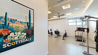 Located in Scottsdale, Arizona, this dedicated fitness room delivers a clean, modern workout environment designed for serious exercise. The space is thoughtfully laid out to accommodate a diverse range of fitness equipment, including a treadmill, recumbent bike, rowing machine, elliptical, weight bench with free weights, and a Life Fitness functional trainer cable machine — providing options for both cardio and strength training.

The room features a coffered tray ceiling accented by warm LED ambient lighting, recessed can lights, and large flush skylight panels that contribute to a bright, energizing atmosphere. A ceiling fan centered within the tray keeps air circulating throughout workouts. The walls are finished in a crisp, clean white, and a large wall-mounted mirror on one side visually expands the space while allowing users to monitor their form.

Durable speckled epoxy flooring covers the entire room, offering both easy maintenance and a polished, professional aesthetic well-suited to a gym environment. A window on the exterior wall allows natural light to filter in, reinforcing the open, airy feel. A vibrant Scottsdale-themed poster near the entrance adds a local character and welcoming touch. Climate control is handled via ceiling-mounted HVAC vents, ensuring comfort year-round in the Arizona heat.