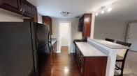 This single-floor space offers a thoughtfully designed interior with a strong emphasis on classic, warm finishes throughout. The kitchen is the centerpiece of the home, styled in a galley layout that maximizes functionality and flow. Rich, dark cherry wood cabinetry lines both sides of the kitchen, providing generous storage from floor to ceiling. The countertops feature a granite-look surface that complements the deep tones of the cabinetry, while the full suite of black appliances — including a refrigerator, gas range, and built-in microwave — adds a sleek, cohesive aesthetic. Track lighting and a flush-mount ceiling fixture ensure the kitchen is well-lit throughout the day. The rich hardwood flooring runs the length of the space, adding warmth and continuity between areas. A doorway at the end of the kitchen leads to an adjoining room, hinting at a well-connected and practical floor plan. Neutral gray-toned walls serve as a clean backdrop that allows the natural richness of the wood elements to take center stage. Overall, the space exudes a timeless, functional charm suited for those who appreciate classic design with modern conveniences.