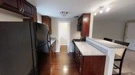 This single-floor space offers a thoughtfully designed interior with a strong emphasis on classic, warm finishes throughout. The kitchen is the centerpiece of the home, styled in a galley layout that maximizes functionality and flow. Rich, dark cherry wood cabinetry lines both sides of the kitchen, providing generous storage from floor to ceiling. The countertops feature a granite-look surface that complements the deep tones of the cabinetry, while the full suite of black appliances — including a refrigerator, gas range, and built-in microwave — adds a sleek, cohesive aesthetic. Track lighting and a flush-mount ceiling fixture ensure the kitchen is well-lit throughout the day. The rich hardwood flooring runs the length of the space, adding warmth and continuity between areas. A doorway at the end of the kitchen leads to an adjoining room, hinting at a well-connected and practical floor plan. Neutral gray-toned walls serve as a clean backdrop that allows the natural richness of the wood elements to take center stage. Overall, the space exudes a timeless, functional charm suited for those who appreciate classic design with modern conveniences.