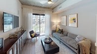 This single-floor residence showcases a thoughtfully designed contemporary interior with industrial-inspired architectural details, including exposed concrete ceilings that lend a distinctive urban character throughout the space. The open-concept kitchen is a standout feature, equipped with a full suite of stainless steel appliances — including a French door refrigerator, electric range with microwave above, and dishwasher. Flat-front, warm-toned wood cabinetry extends floor to ceiling, complemented by a neutral tile backsplash, white quartz countertops, and under-cabinet lighting. A granite-topped kitchen island provides additional prep and casual dining space, with pendant lighting above. Wood-look flooring flows through the kitchen and living areas, adding warmth to the industrial aesthetic. The living room opens to a private balcony through floor-to-ceiling sliding glass doors, inviting natural light and outdoor views. The bedroom features a dramatic dark accent wall, exposed concrete ceiling, large windows with sheer drapery, and ample space for a dresser and seating. The bathroom is finished with large-format light gray floor tiles and marble-style wall tile surrounding a soaking tub, creating a clean, spa-like atmosphere.