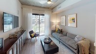 This single-floor residence showcases a thoughtfully designed contemporary interior with industrial-inspired architectural details, including exposed concrete ceilings that lend a distinctive urban character throughout the space. The open-concept kitchen is a standout feature, equipped with a full suite of stainless steel appliances — including a French door refrigerator, electric range with microwave above, and dishwasher. Flat-front, warm-toned wood cabinetry extends floor to ceiling, complemented by a neutral tile backsplash, white quartz countertops, and under-cabinet lighting. A granite-topped kitchen island provides additional prep and casual dining space, with pendant lighting above. Wood-look flooring flows through the kitchen and living areas, adding warmth to the industrial aesthetic. The living room opens to a private balcony through floor-to-ceiling sliding glass doors, inviting natural light and outdoor views. The bedroom features a dramatic dark accent wall, exposed concrete ceiling, large windows with sheer drapery, and ample space for a dresser and seating. The bathroom is finished with large-format light gray floor tiles and marble-style wall tile surrounding a soaking tub, creating a clean, spa-like atmosphere.